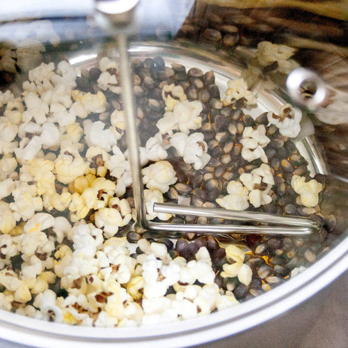 Partially popped popcorn kernels inside a stainless steel stovetop popper with a hand crank stirring arm, handcrafted for Kitchen & Food, Cookware, and Popcorn collections.