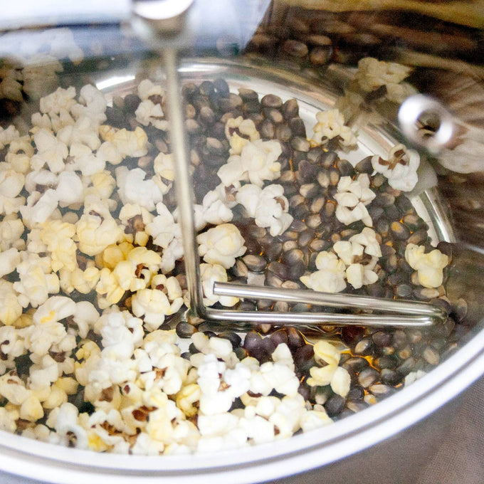 Partially popped popcorn kernels inside a stainless steel stovetop popper with a hand crank stirring arm, handcrafted for Kitchen & Food, Cookware, and Popcorn collections.