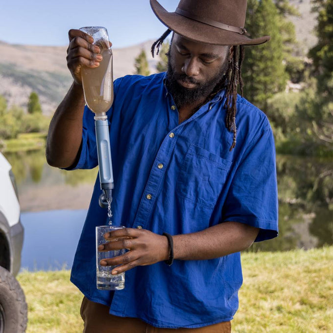 Man wearing a wide-brimmed hat and blue shirt outdoors filtering muddy water through a portable blue water filter straw into a clear glass near a lake in a natural camping setting