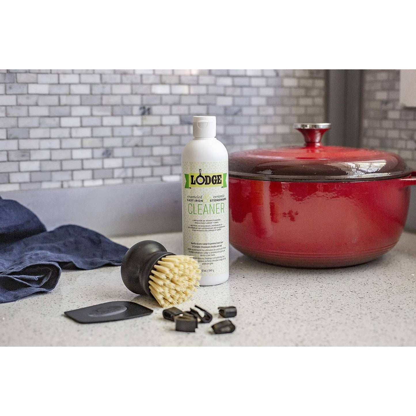 Biodegradable cleaner bottle, polycarbonate pan scraper, brush with stiff bristles, and black cleaning clips displayed on kitchen counter next to a red enameled cast iron pot, suitable for Cast Iron and Kitchen & Food collections.