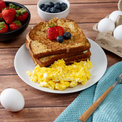 Plate of fluffy scrambled eggs and two slices of French toast garnished with fresh strawberries and blueberries on a wooden table with whole eggs, a bowl of strawberries, and a bowl of blueberries nearby, evoking a comforting breakfast setting suitable for camping and emergency food supplies.
