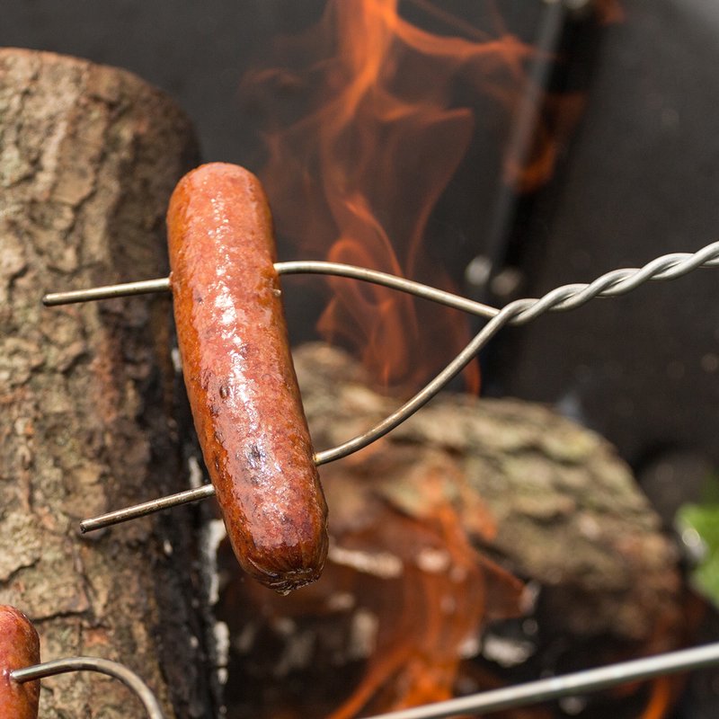 Heavy-duty, galvanized steel roasting stick with a hot dog secured by two prongs over an open campfire, featuring an extra-long wooden handle, great for Camping and USA Made collections.