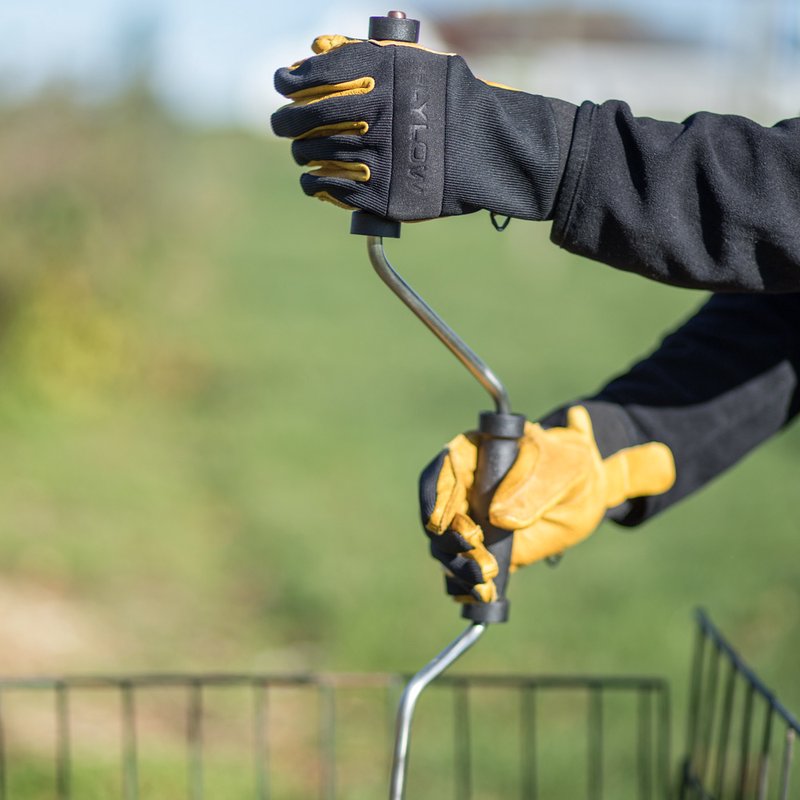 Gloved hands turning a stainless steel compost crank with a curved handle, used for aerating and mixing compost bins in outdoor garden and farm settings.