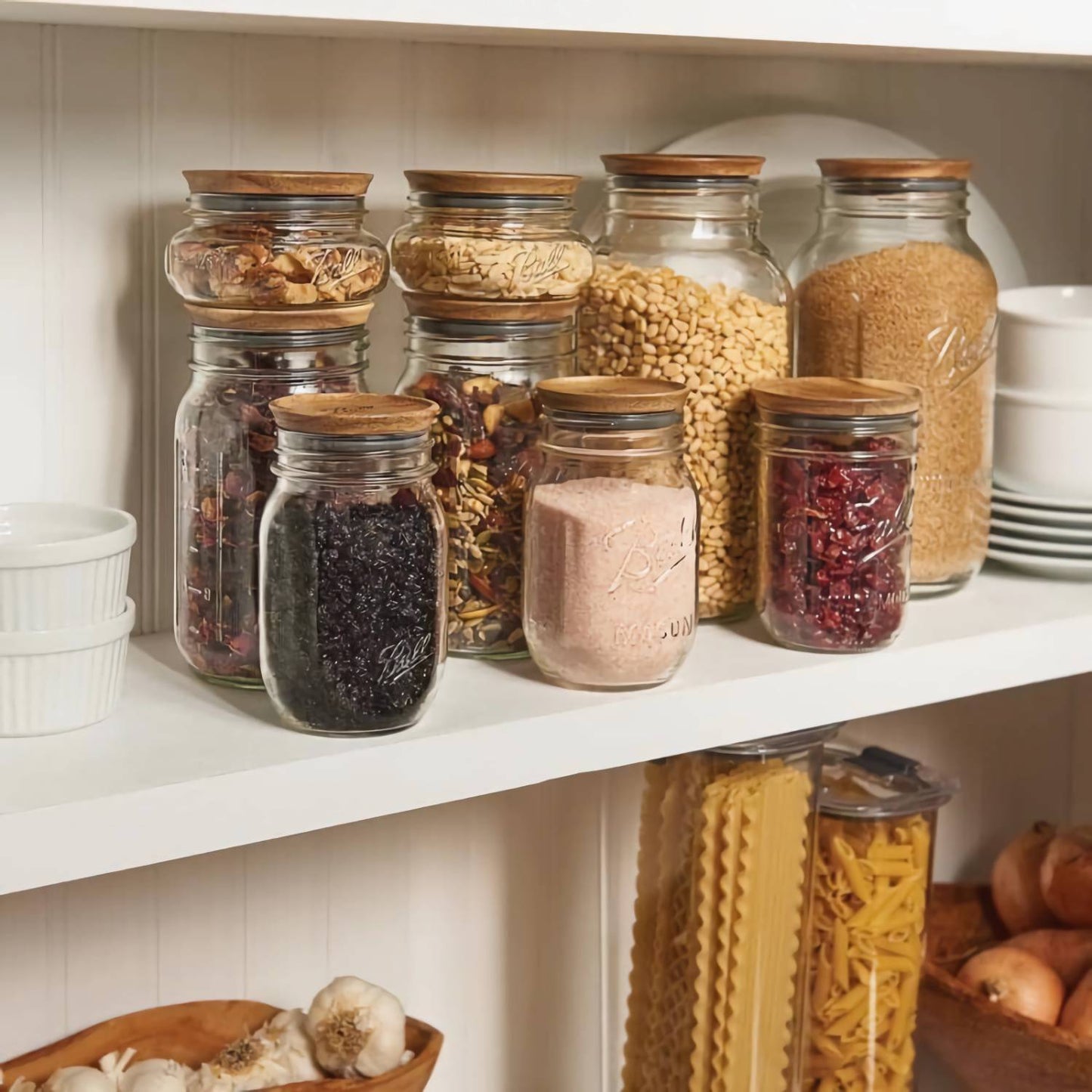 Set of clear glass storage jars with airtight acacia wooden lids in various sizes, filled with dry pantry goods and organized on a white shelf, featuring a warm kitchen and food storage scene.