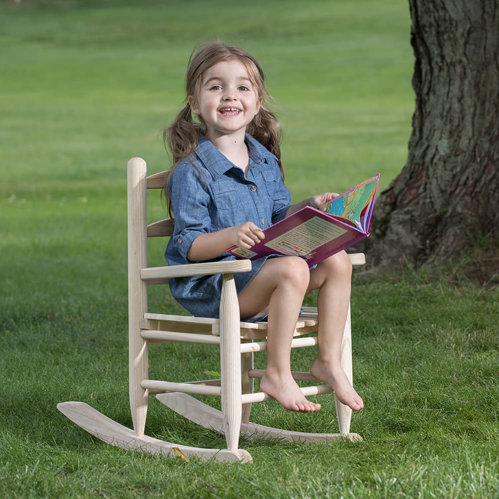 Smiling child reading a colorful book while sitting on a handcrafted Amish-made unfinished ash wooden rocker outdoors on green grass, part of children's toys and wooden toys collections.