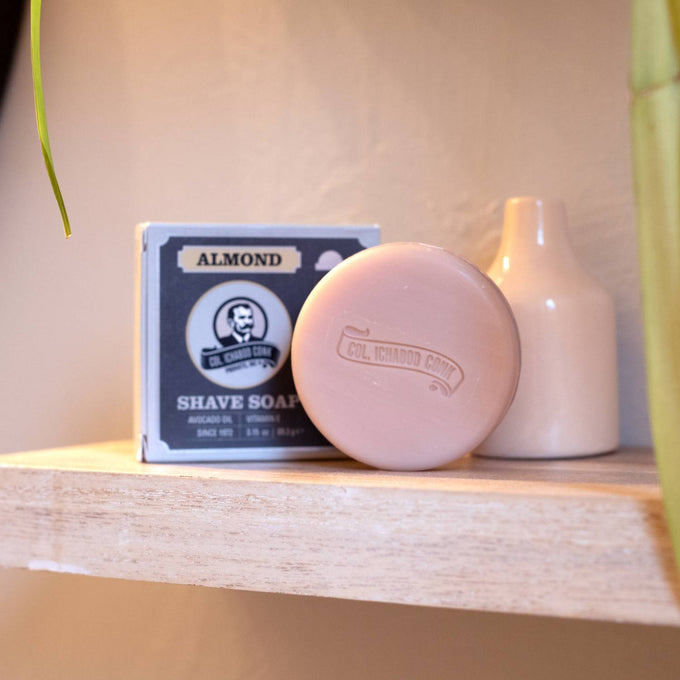 Round almond-scented shaving soap bar with embossed logo, displayed next to its boxed packaging on a wooden shelf, featured in Health & Wellness and Grooming collections.