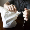 Hands tightening the drawstring on a white reusable fabric bag made to keep leafy greens fresh longer, featured in Kitchen & Food, Storage and Serving, and Best Sellers collections.