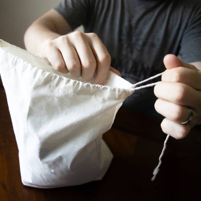 Hands tightening the drawstring on a white reusable fabric bag made to keep leafy greens fresh longer, featured in Kitchen & Food, Storage and Serving, and Best Sellers collections.