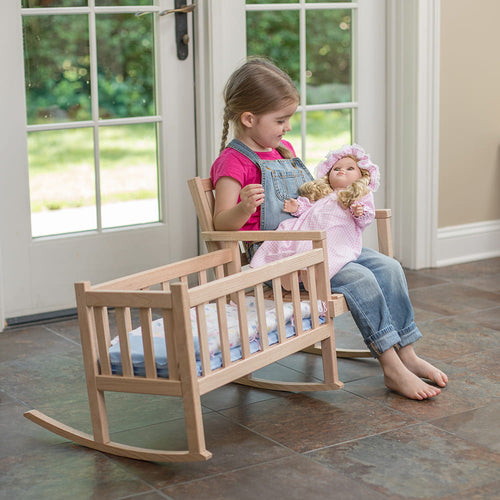 Young child sitting barefoot on a handcrafted unfinished oak doll rocker, holding a doll dressed in pink, next to a matching wooden doll cradle, set indoors near glass doors, showcasing Amish-made children's toys from USA Made, Amish Made, and Toys, Books & Games collections.