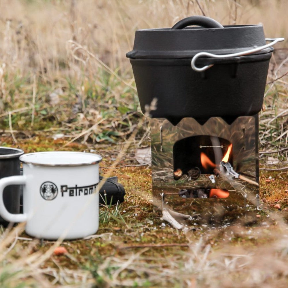 Black cast iron Dutch oven with flat base cooking over an open flame on a stainless steel portable stove outdoors, accompanied by a white enamel camping mug, featured in Kitchen & Food, Cookware, Cast Iron, Best Sellers, and Promo Eligible collections.
