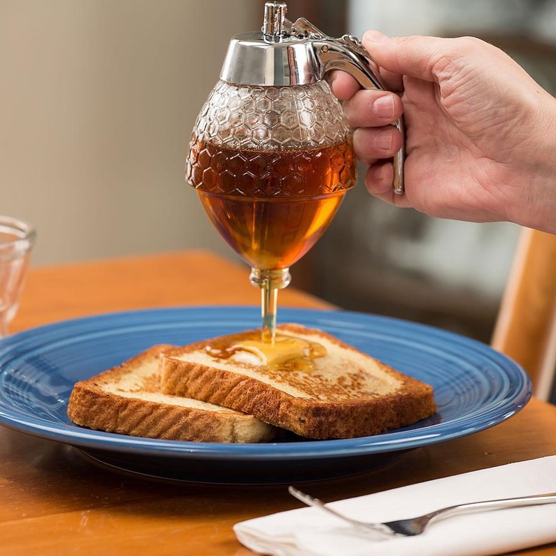 Glass dispenser with textured honeycomb design and metal lever, pouring honey onto toast on a blue plate, ideal for Kitchen & Food, Storage and Serving, Best Sellers, and Promo Eligible collections.