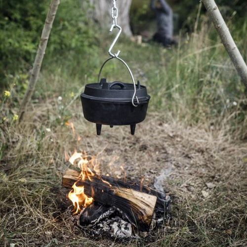 Handcrafted cast iron Dutch oven with three legs and a bail handle suspended over an open wood fire in a natural outdoor setting, perfect for Kitchen & Food, Cookware, Cast Iron, Best Sellers, and Promo Eligible collections.