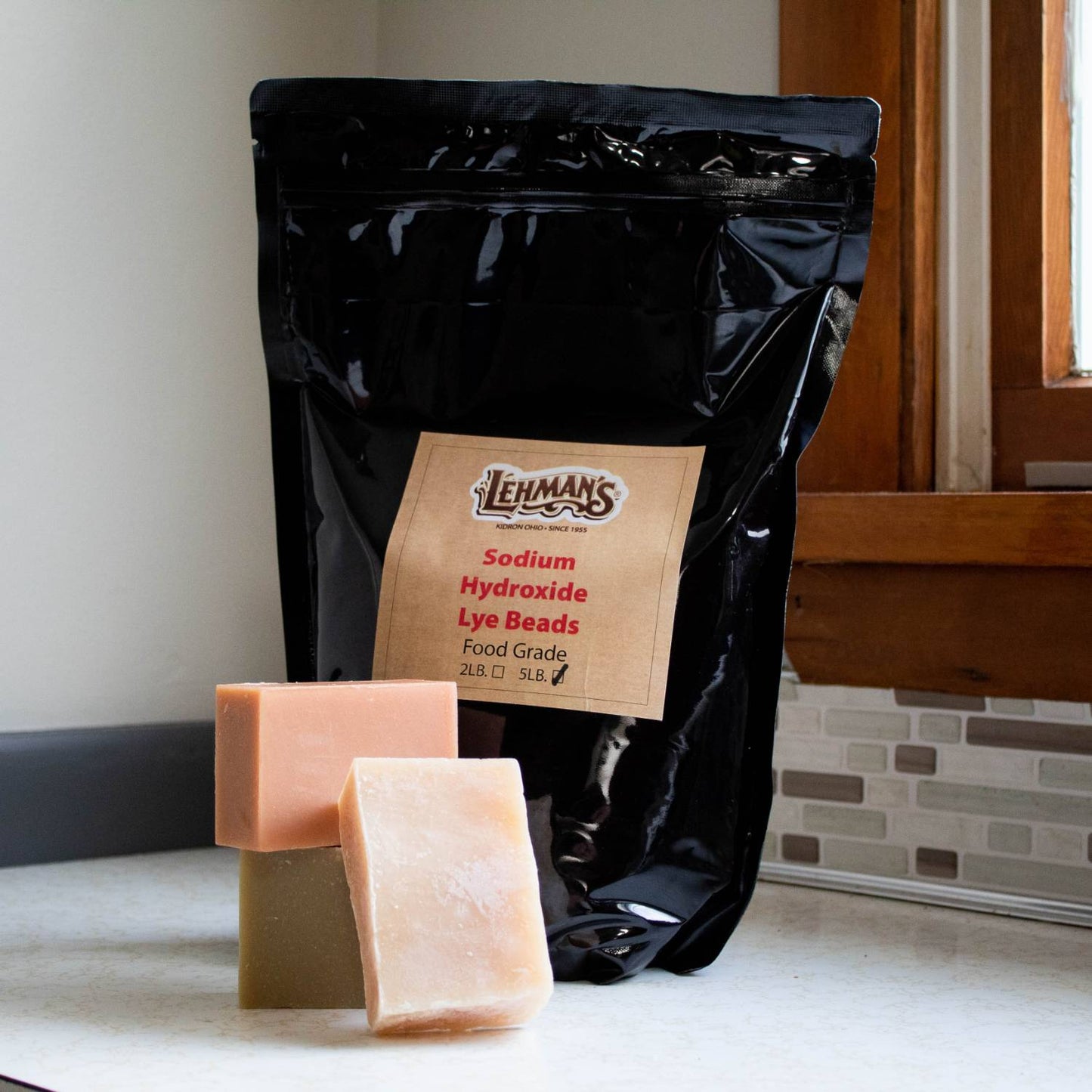 Black resealable pouch of high-grade sodium hydroxide lye beads with three handmade soap bars displayed on a countertop, part of health and wellness and soapmaking supplies collections.
