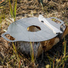 Oiled steel griddle plate with dual handles and a central hole, resting on a birch log in outdoor grass, ideal for versatile grilling in camping, farm, and stove collections.