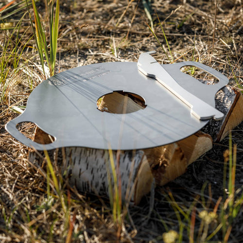 Oiled steel griddle plate with dual handles and a central hole, resting on a birch log in outdoor grass, ideal for versatile grilling in camping, farm, and stove collections.