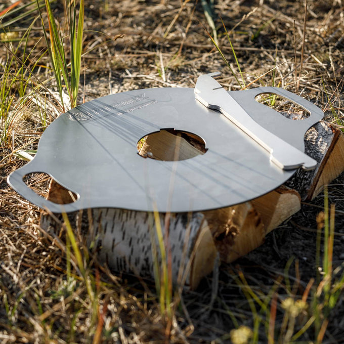 Oiled steel griddle plate with dual handles and a central hole, resting on a birch log in outdoor grass, ideal for versatile grilling in camping, farm, and stove collections.