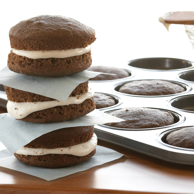 Stack of three chocolate whoopie pies with creamy white filling separated by parchment paper next to a metal baking pan with freshly baked cakes, ideal for Kitchen & Food and Baking Supplies collections.