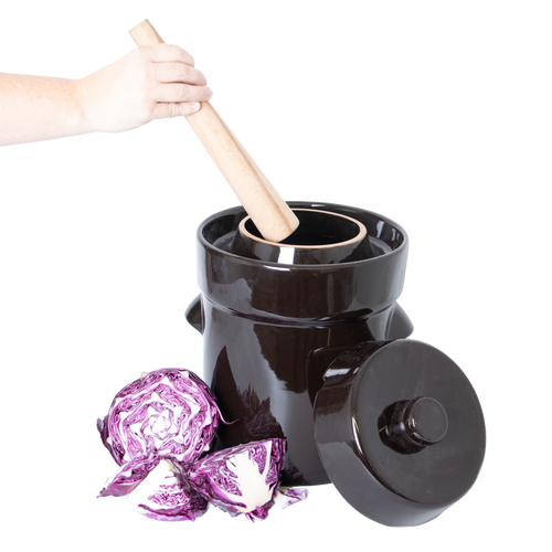 Handheld wooden stomper pressing into a dark ceramic crock with lid resting beside it, surrounded by fresh purple cabbage, for kitchen, food preservation, and fermenting use.