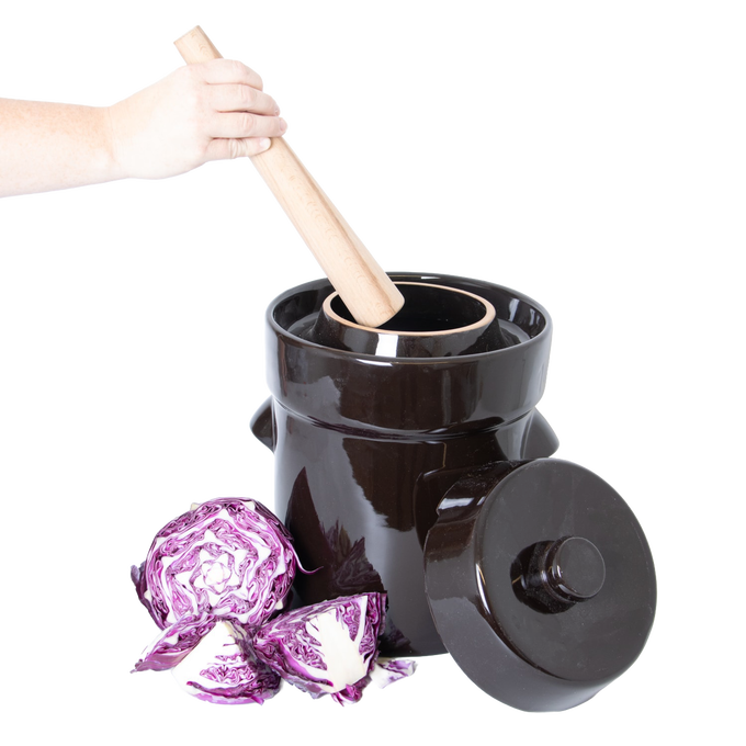 Handheld wooden stomper pressing into a dark ceramic crock with lid resting beside it, surrounded by fresh purple cabbage, for kitchen, food preservation, and fermenting use.
