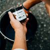 Person holding a small container of Petromax Care Conditioner for cast and wrought iron, positioned above a black cast iron pot, set outdoors on a textured ground surface, suitable for Kitchen & Food, Cookware, Cast Iron, Best Sellers, and Promo Eligible collections.