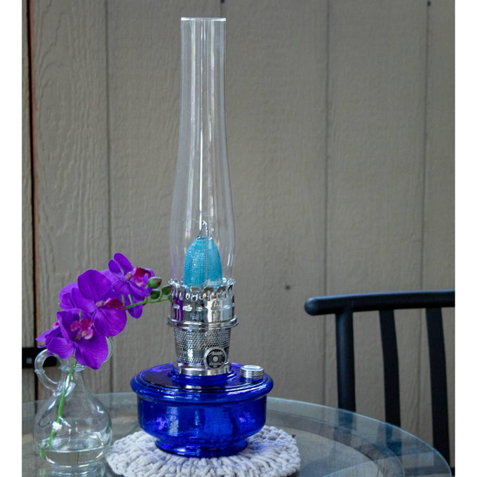 Handcrafted Aladdin Genie III oil lamp with deep blue dimpled glass base and gleaming brass burner, displayed on glass table with purple orchid, ideal for Farm, Garden & Tool and Lamps & Lighting collections.
