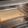 Large silicone baking mat with a tan center and orange border placed on a metal baking sheet inside a home oven, ideal for Kitchen & Food, Cookware, Baking Supplies, and Best Sellers collections.