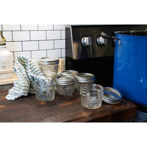 Set of clear quilted glass jars with silver lids arranged on wooden counter next to stove and blue pot, designed for food preservation and canning, made in USA, part of Kitchen & Food and Canning collections.