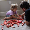 Young child and adult playing together indoors with handcrafted red wooden log pieces from Lehman's Fort Wooden Log Playset, promoting creative indoor play and imagination.