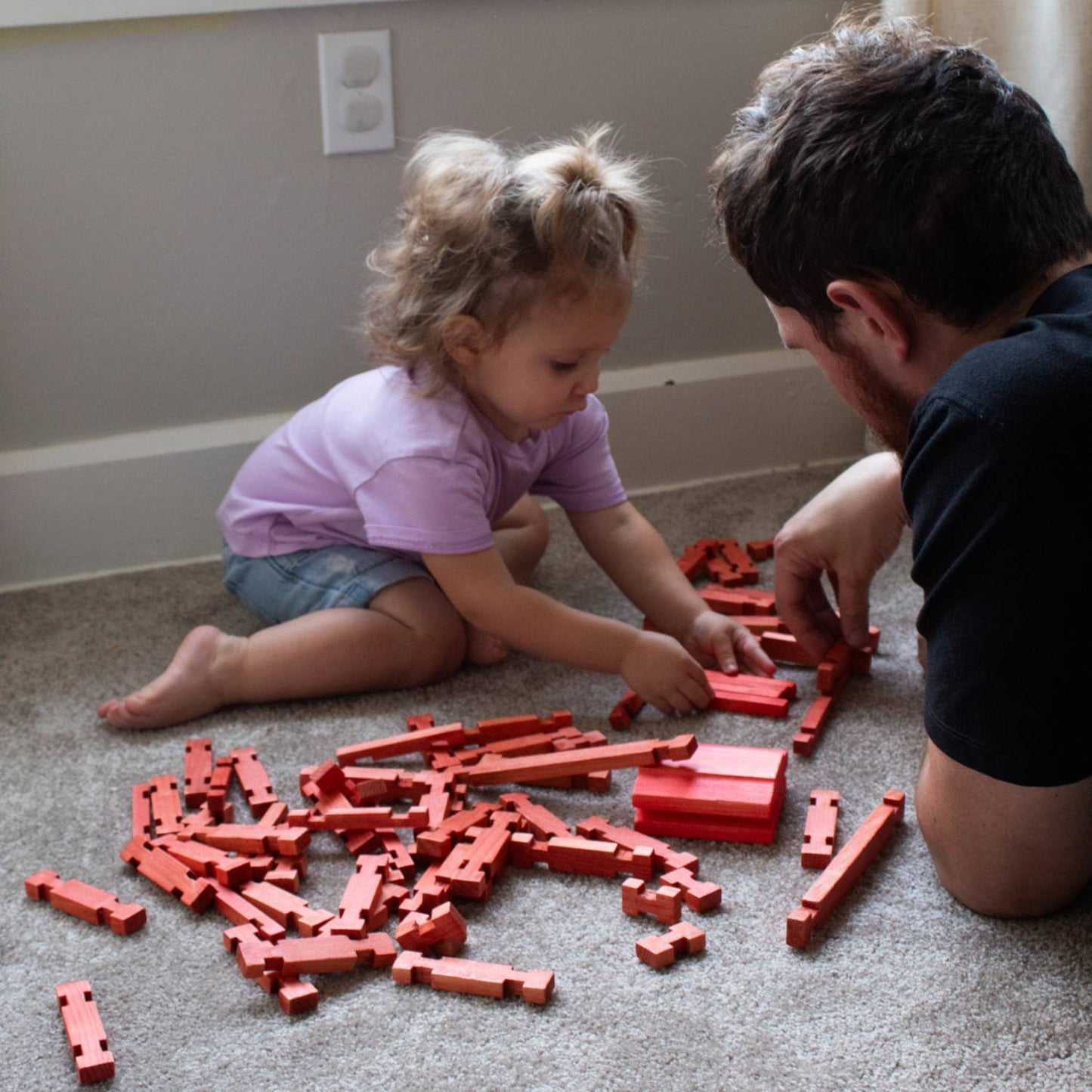 Young child and adult playing together indoors with handcrafted red wooden log pieces from Lehman's Fort Wooden Log Playset, promoting creative indoor play and imagination.