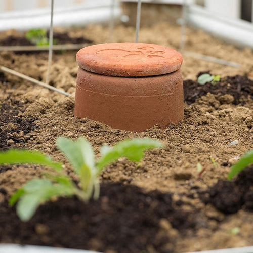 Unglazed terra cotta watering pot partially buried in garden soil, handcrafted for slow water release to conserve moisture in small beds and planters, featured in Farm, Garden & Tool, Garden Accessories, Lawncare, Best Sellers, and Promo Eligible collections.