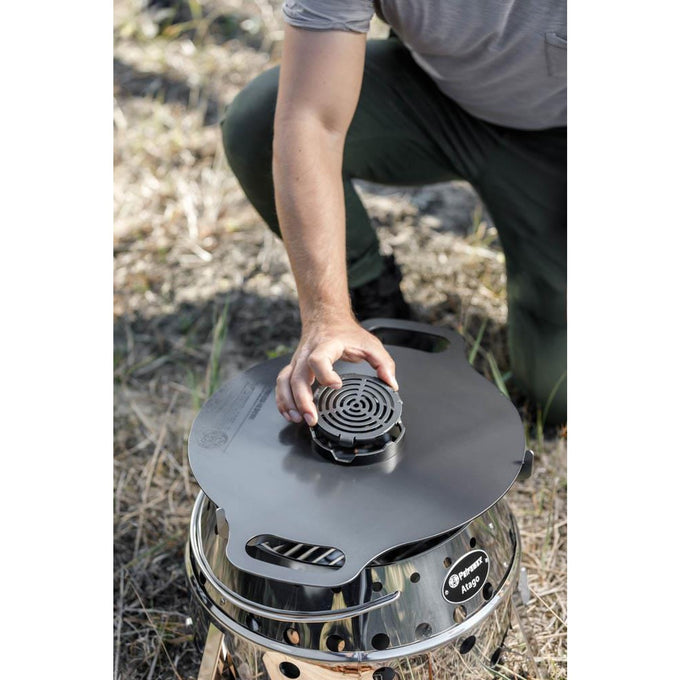 Person placing a round metal insert with a concentric ring design into the center opening of a black griddle plate set on a shiny outdoor cooking stove, suited for Farm, Garden & Tool, Grilling, and Camping collections.
