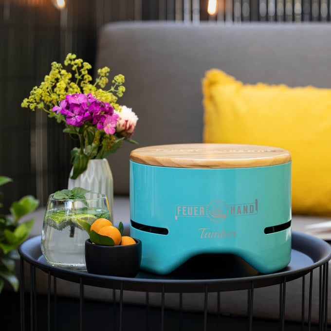 Handcrafted turquoise tabletop charcoal grill with a smooth wooden lid, displayed on a round metal outdoor table alongside fresh flowers, fruit, and a refreshing drink, ideal for Farm, Garden & Tool, Camping, and Grilling collections.