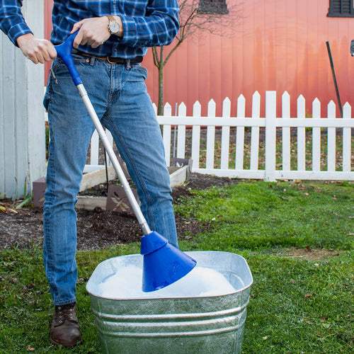 Person using a blue-handled collapsible hand washer plunger in a galvanized metal tub filled with soapy water outdoors near a white picket fence, part of laundry supplies for Home & Decor and Washers and Wash Day Accessories collections.