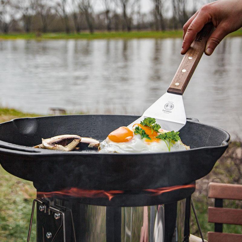 Handcrafted cast iron skillet with two side handles cooking sunny-side-up eggs garnished with fresh parsley over an outdoor camping stove, suitable for farm, kitchen, and cookware use.