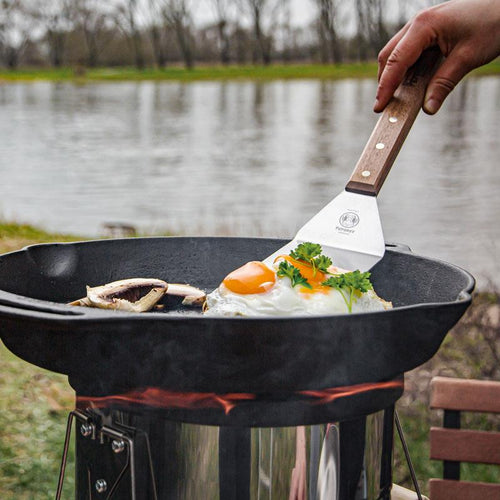 Handcrafted cast iron skillet with two side handles cooking sunny-side-up eggs garnished with fresh parsley over an outdoor camping stove, suitable for farm, kitchen, and cookware use.