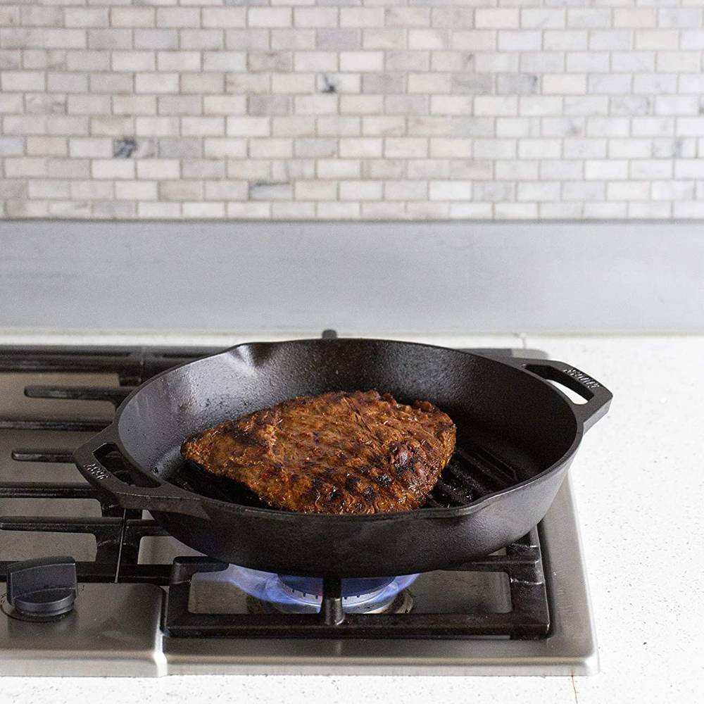 Seasoned cast iron grill pan with dual handles heating on a gas stove, searing a piece of meat, showcasing durable cookware from USA made kitchen and cast iron collections.