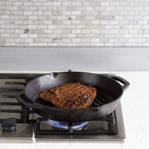 Seasoned cast iron grill pan with dual handles heating on a gas stove, searing a piece of meat, showcasing durable cookware from USA made kitchen and cast iron collections.