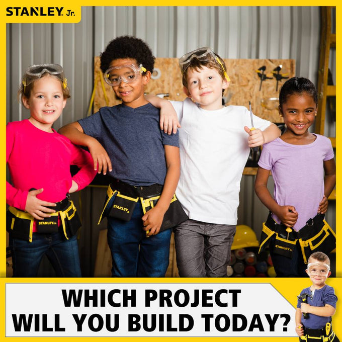 Four children wearing safety goggles and Stanley-branded tool belts pose confidently in a workshop setting, promoting creativity and hands-on learning in Toys, Books & Games and Arts and Crafts collections.