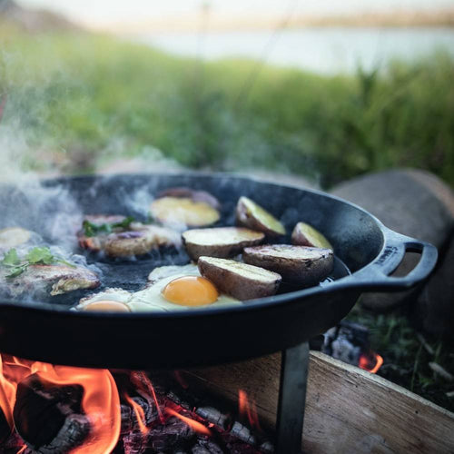 Cast iron skillet with two side handles cooking eggs, potatoes, and vegetables over open fire outdoors, perfect for camping, farm, kitchen, and cookware collections.