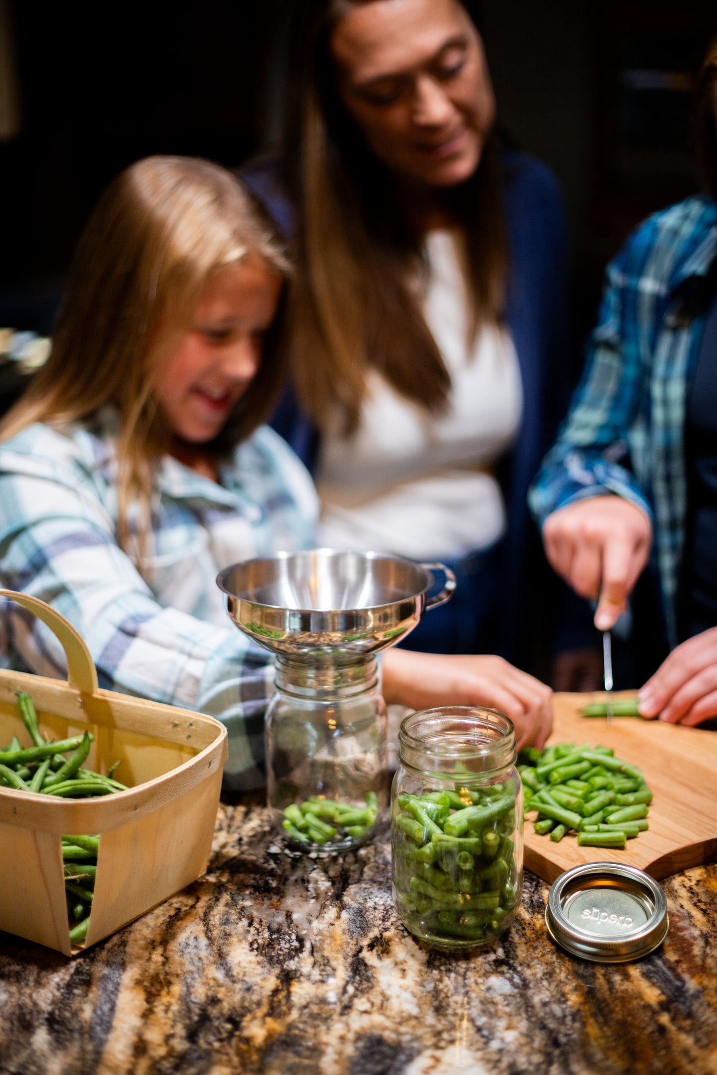 Family preparing fresh green beans for preserving using a stainless steel funnel and glass jars on a granite countertop, part of home and garden how-to books and best sellers collections.