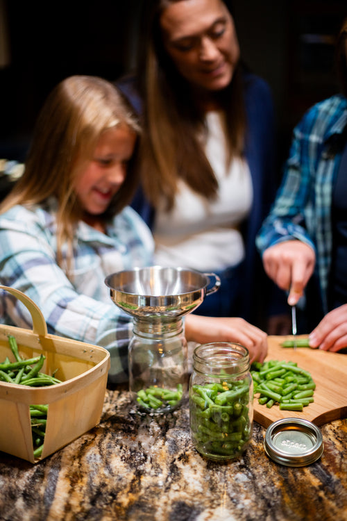 Family preparing fresh green beans for preserving using a stainless steel funnel and glass jars on a granite countertop, part of home and garden how-to books and best sellers collections.