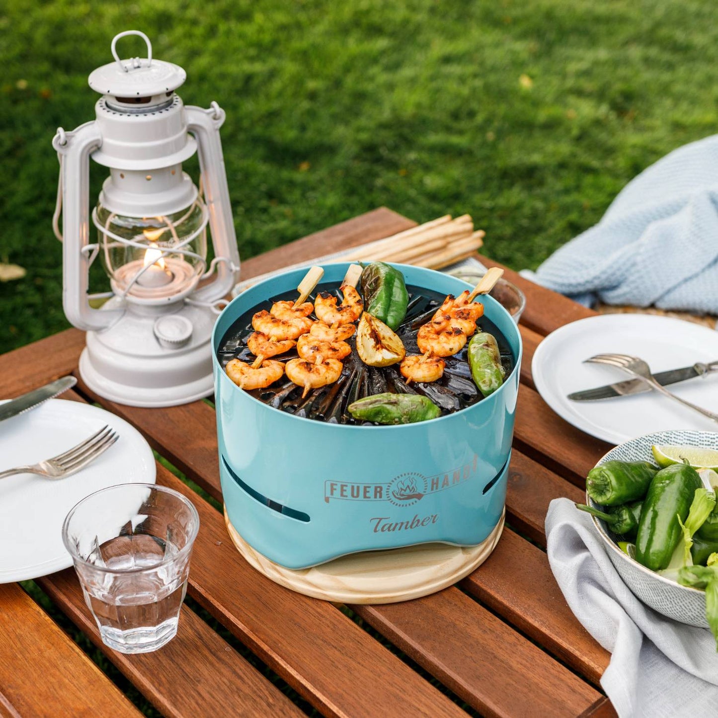 Handcrafted blue tabletop charcoal grill cooking shrimp skewers and charred peppers, set on a wooden outdoor table with a white lantern and simple dining setup, ideal for Farm, Garden & Tool, Camping, and Grilling collections.