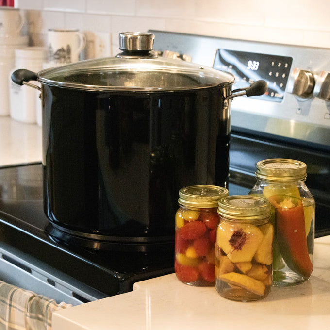 Large black water bath canner with glass lid featuring built-in temperature gauge on stove next to three sealed jars of preserved fruits and vegetables, ideal for Kitchen & Food, Food Preservation, and Canning collections.