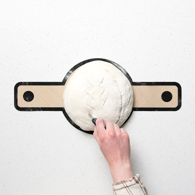 Hand scoring a round loaf of sourdough dough on a wooden bread shaping board with black handles, part of a handcrafted baking set for Kitchen & Food and Bread Making Supplies collections.