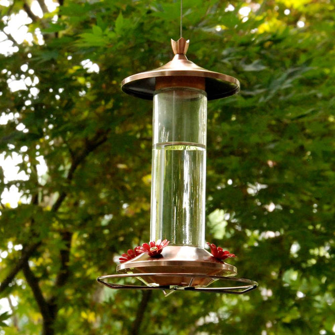 Hanging clear glass feeder with brushed copper base and metal top, featuring four flower-shaped feeding ports, set against green leafy background in Farm, Garden & Tool and Animal Care collections.
