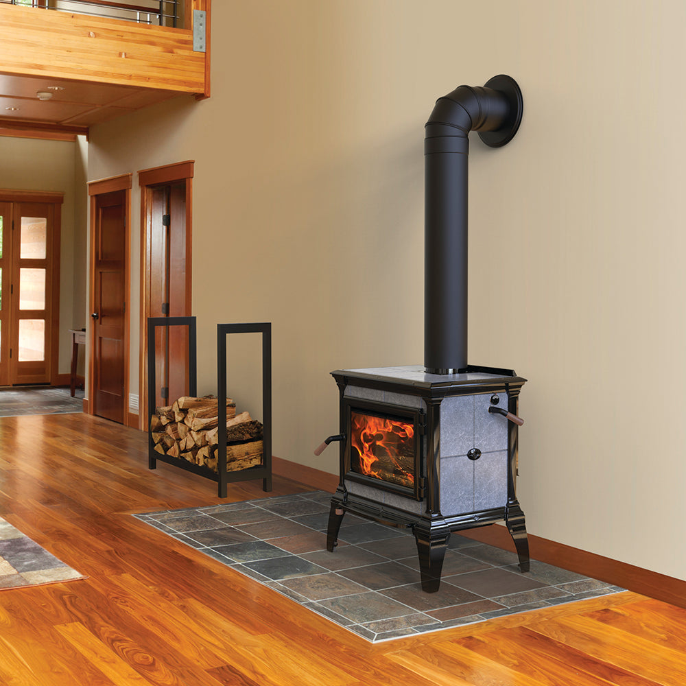 Wood heat stove with a black and stone gray finish, featuring a glass door showing a glowing fire, installed on a tile hearth in a cozy living room setting, part of Stoves and Heatstoves collections.