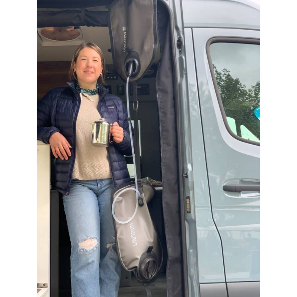 Woman standing in the doorway of a gray camper van holding a metal mug, with a LifeStraw Gravity Water Filter bag hanging nearby, set in an outdoor camping environment.