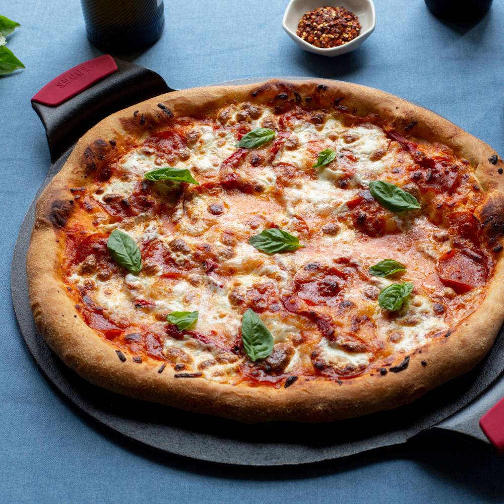 Round cast iron pizza pan with dual handles and red silicone grips holding a freshly baked pepperoni pizza topped with melted cheese and basil, featured in Kitchen & Food, Cookware, Cast Iron, USA Made collections.
