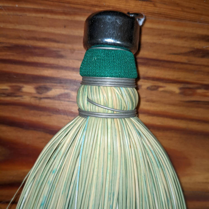 Handcrafted corn whisk broom made in the USA with tightly bound natural corn straw, featuring a metal cap and green fabric band, ideal for household cleaning tasks.