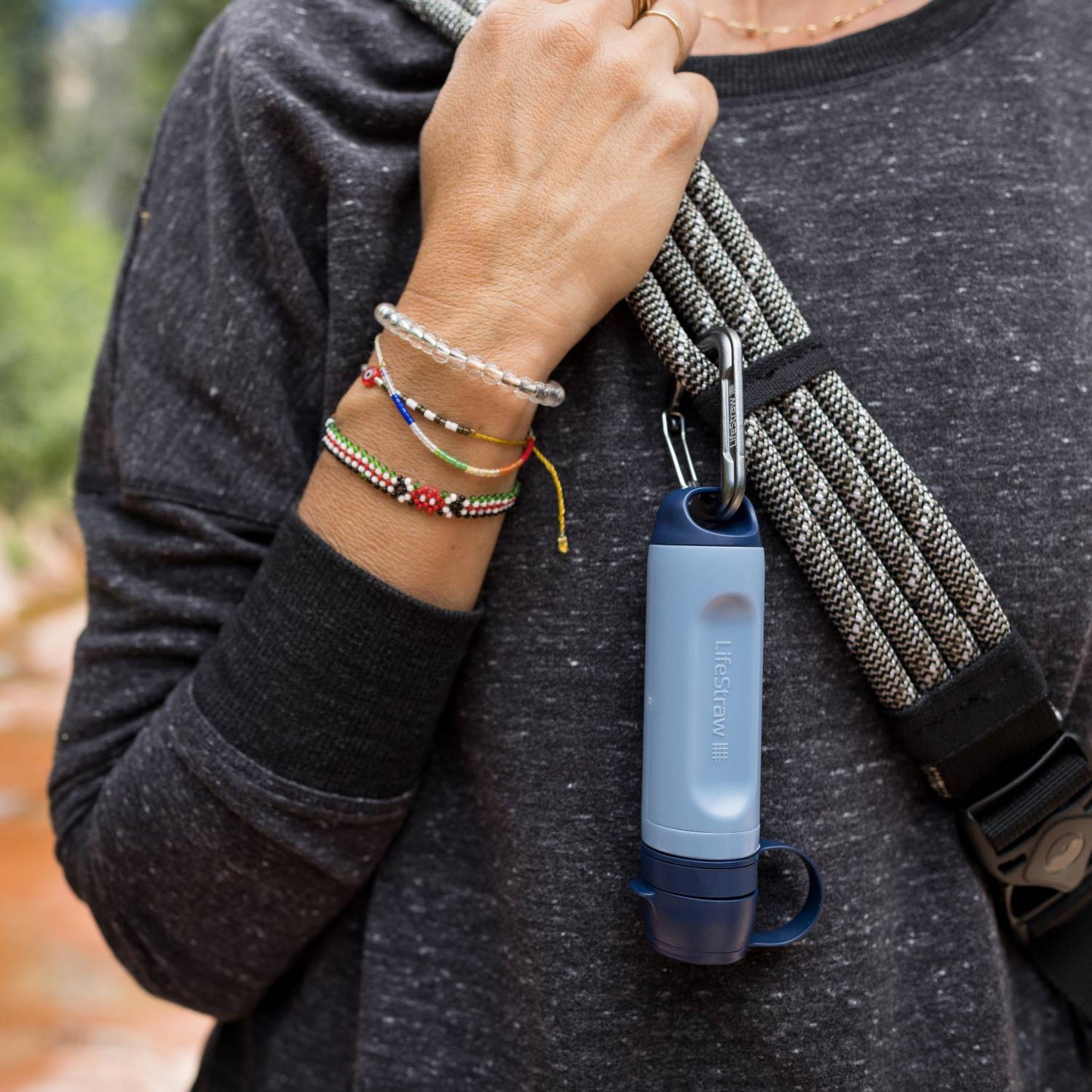 Light blue portable water filter with carabiner clip attached to hiking rope, held by a person wearing a dark long-sleeve shirt and colorful bracelets, suitable for camping and emergency supplies.